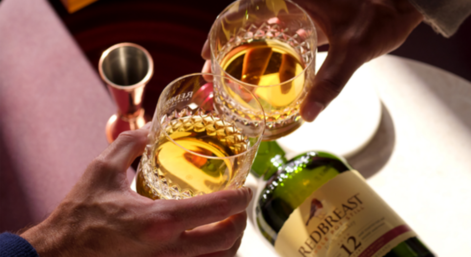 Two handheld whiskey glasses meeting in a cheers motion, with Redbreast 12 bottle on the table underneath.